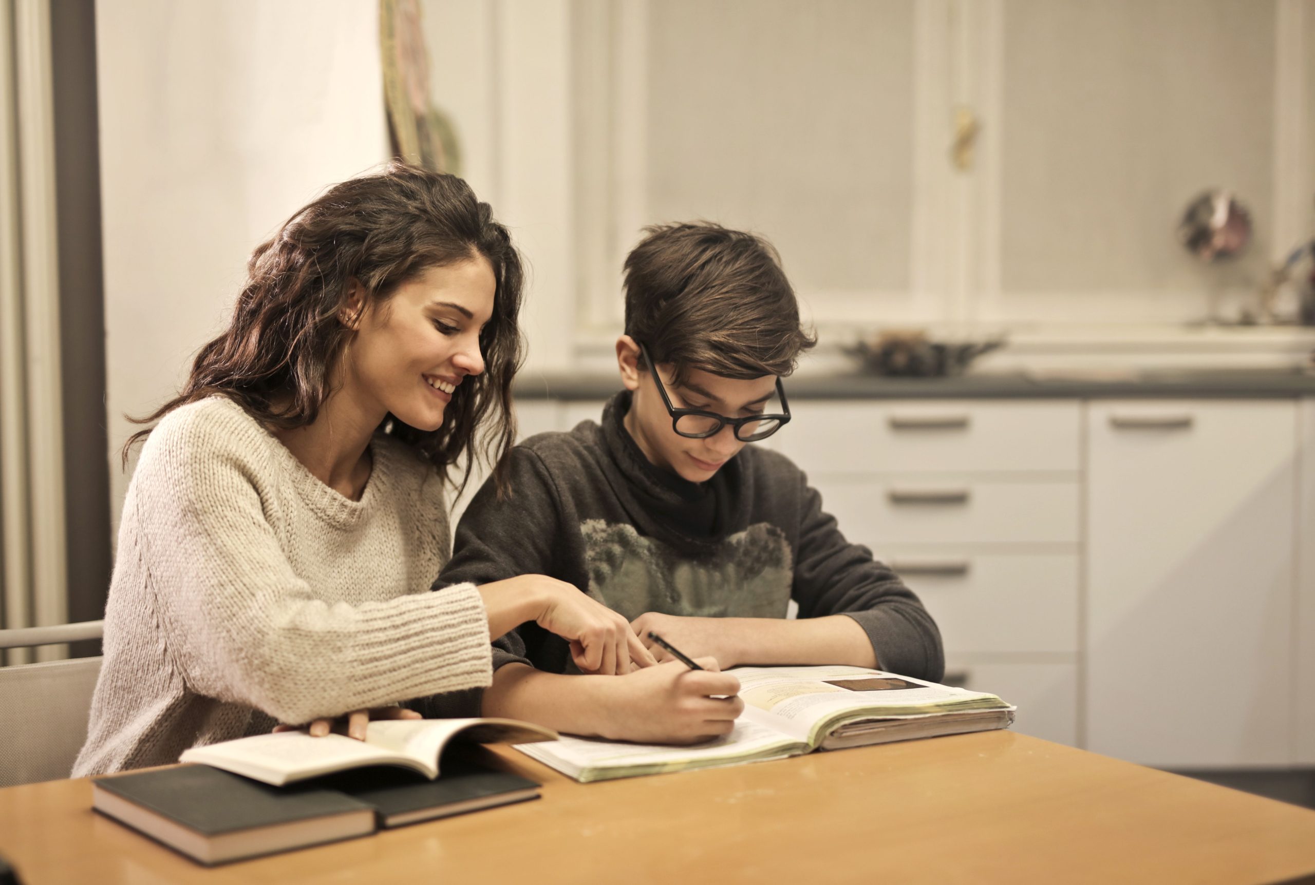 Cheerful young woman helping younger brother with homework while sitting together at table with books at home