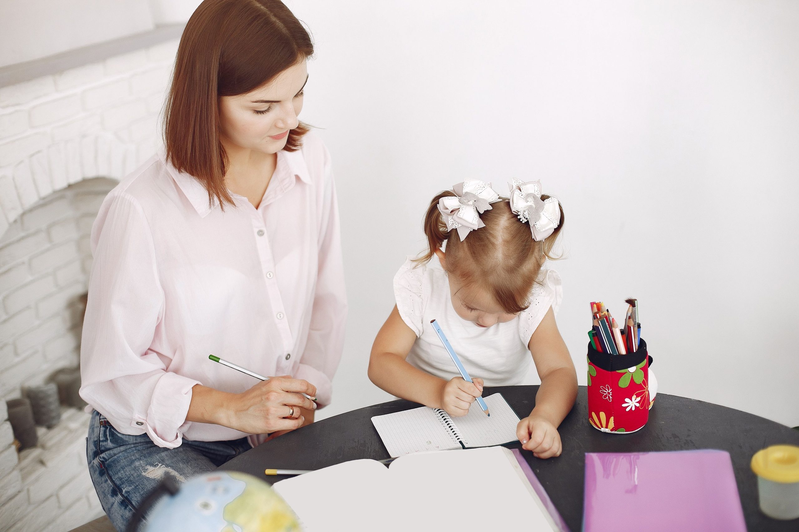 From above small girl drawing with pencil in notepad while mother sitting near at round table